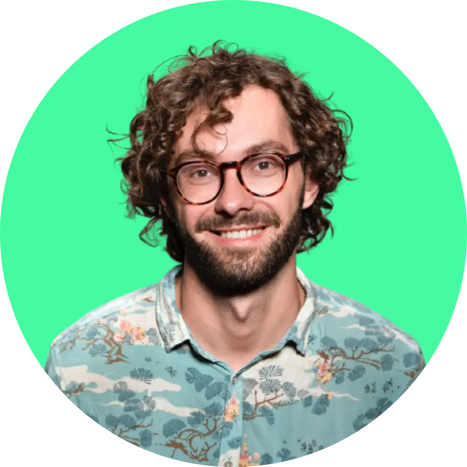 headshot of the founder, a man with a beard and curly brown hair, smiling infront of a green background, wearing glasses and a shirt with tree designs on it